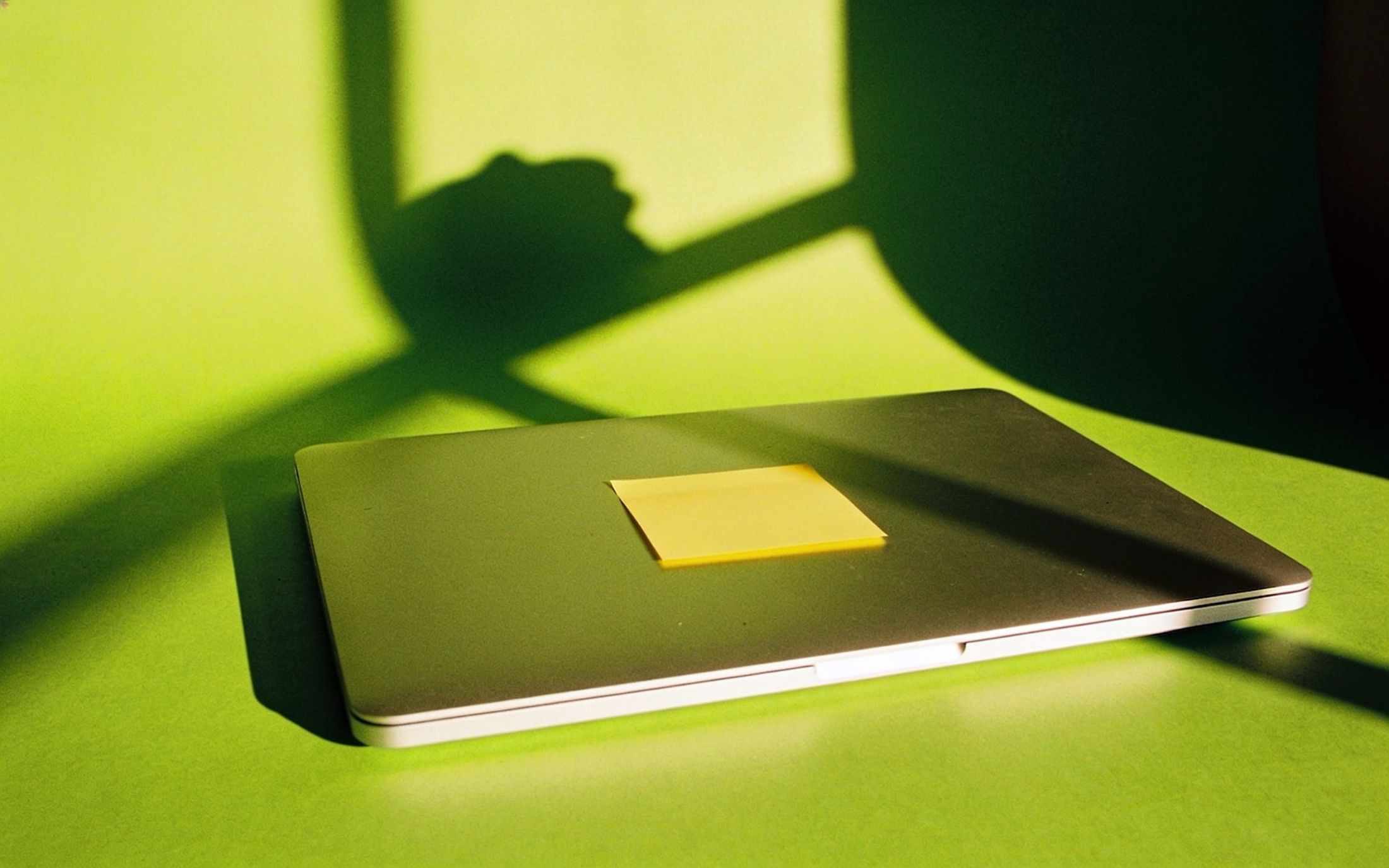 A closed laptop with a yellow sticky note on its lid, long afternoon shadows on lime paper