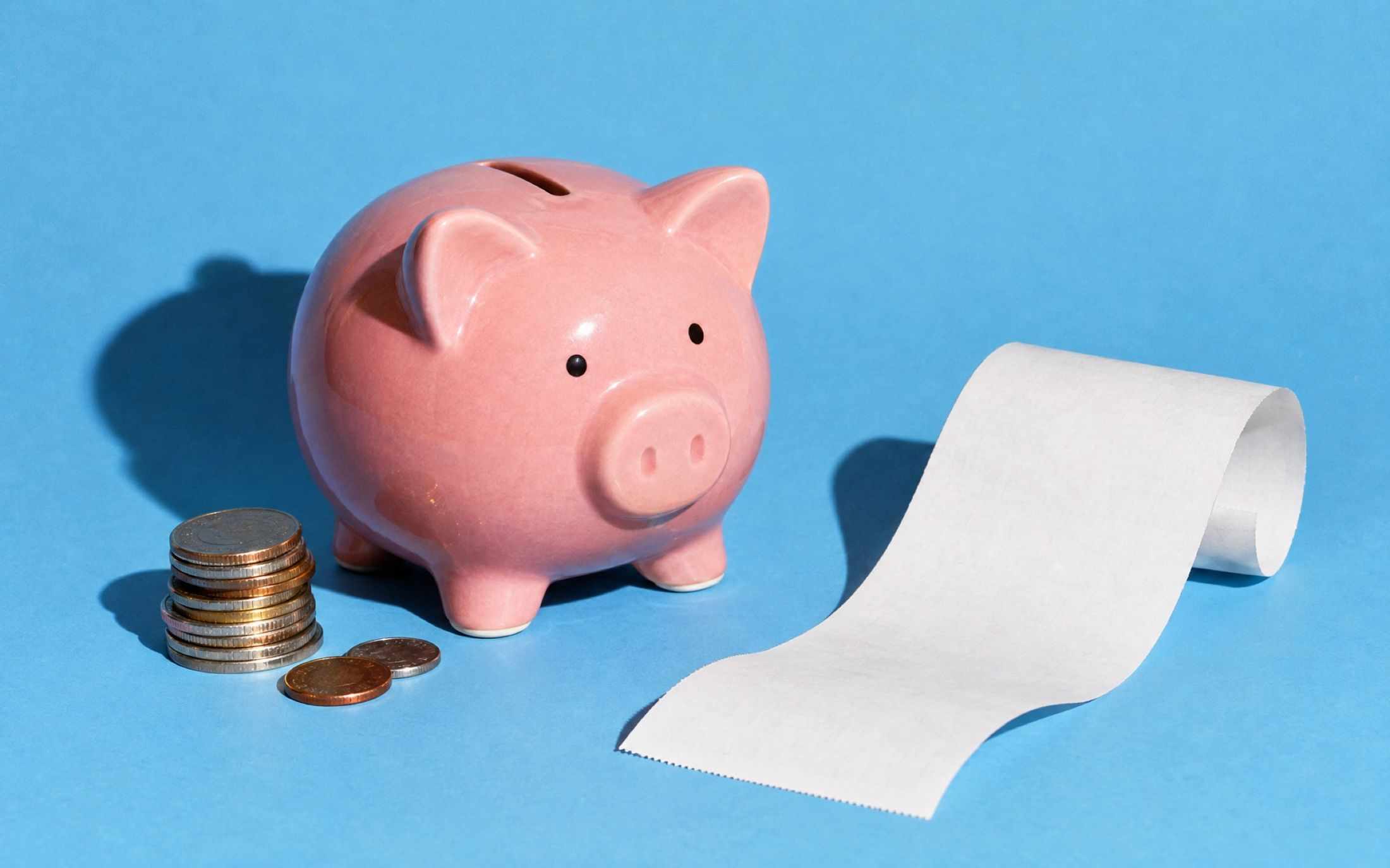 A stack of coins, a pink piggy bank and a curling paper receipt on a sky blue backdrop