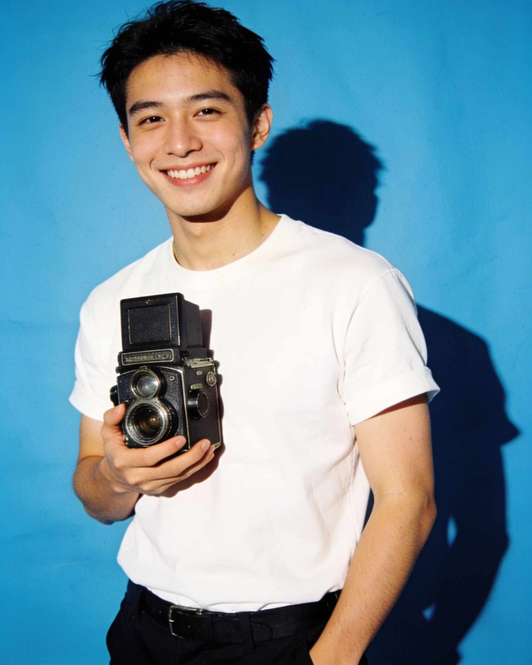 A grinning photographer holding a film camera on a sky blue backdrop