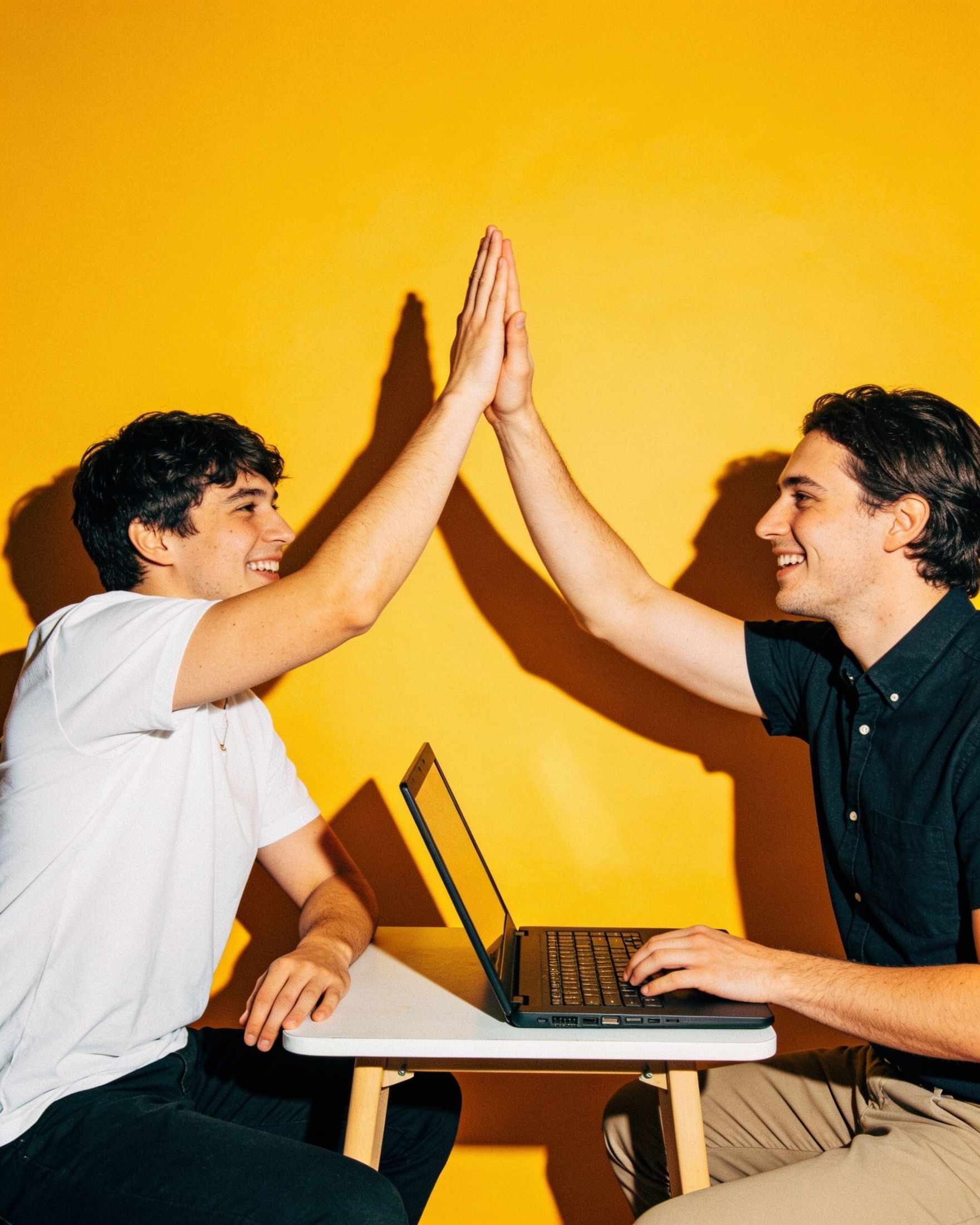 Two colleagues giving a high five over a laptop on a yellow backdrop