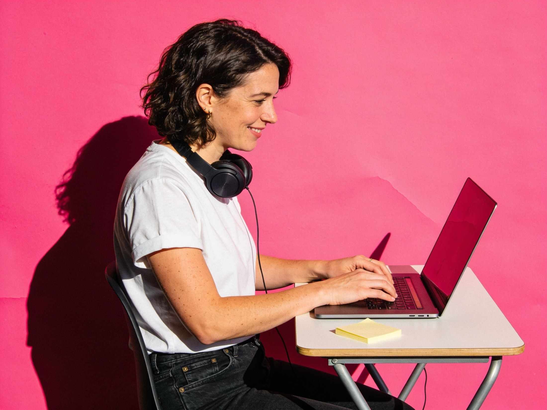 A designer working at a laptop on a pink backdrop, headphones round her neck