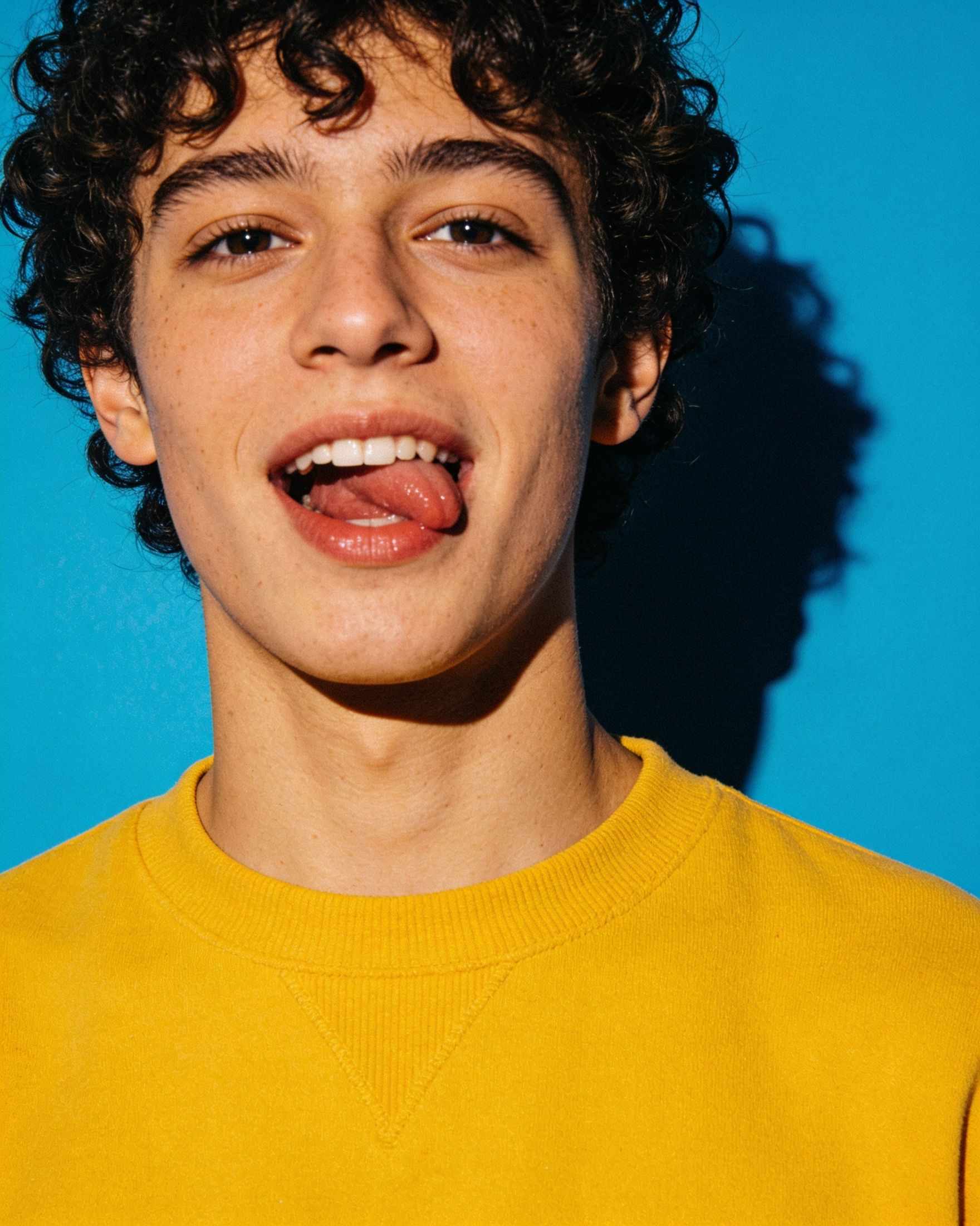 Sami, a young person with curly hair and a yellow jumper, on a sky blue backdrop
