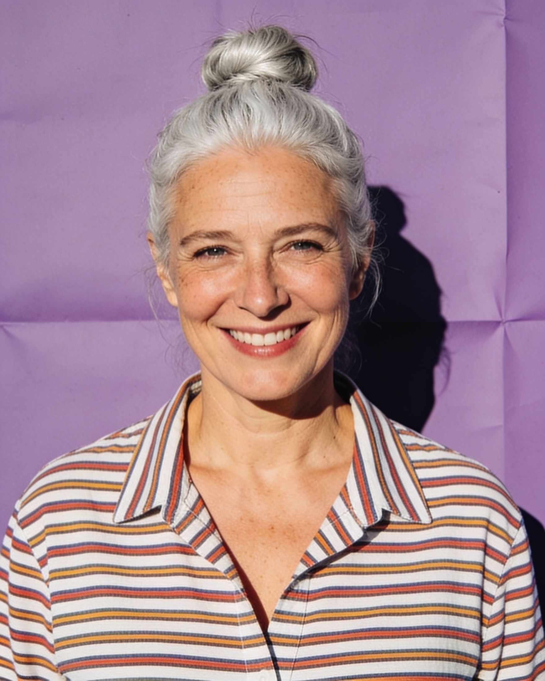 Helen, a woman with silver hair in a bun and a striped shirt, on a lilac backdrop